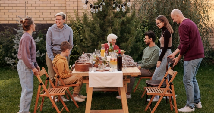 Happy family members talking and sitting down to eat tasty food at big wooden table in backyard in daytime