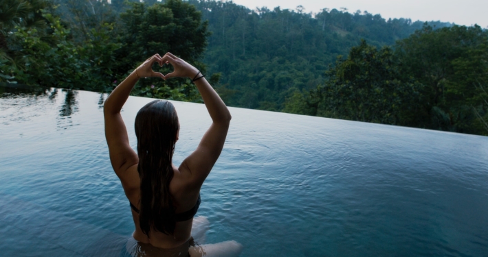 woman in infinity pool making heart hand gesture facing green leafed trees