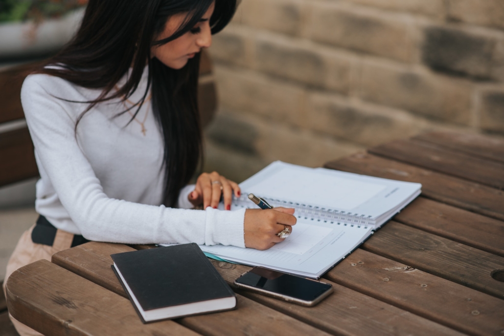 Crop ethnic freelancer writing in notebook at street table