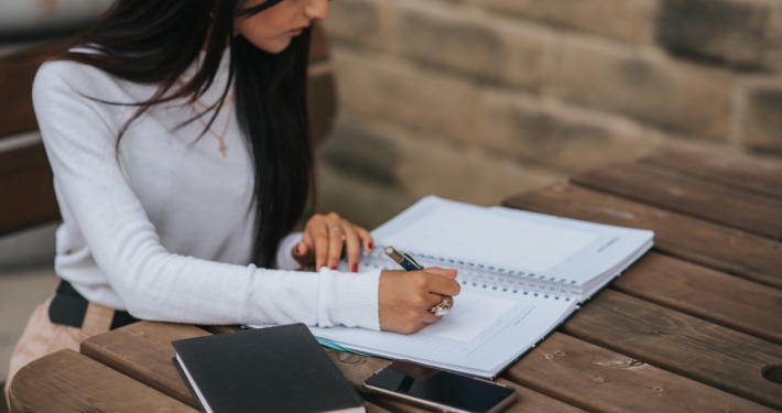 Crop ethnic freelancer writing in notebook at street table