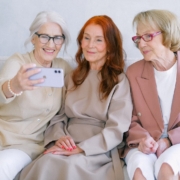 Senior cheerful women using mobile phone and looking at screen while making video call together while resting on sofa together