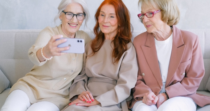 Senior cheerful women using mobile phone and looking at screen while making video call together while resting on sofa together