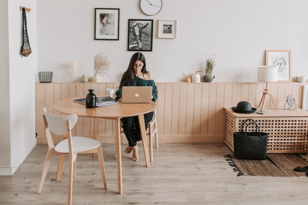 Full length of pensive brunette in eyeglasses sitting at table with legs crossed and working on laptop in cozy creative living room