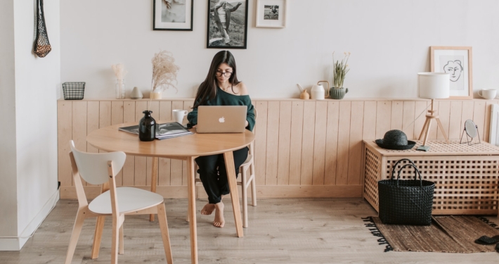 Full length of pensive brunette in eyeglasses sitting at table with legs crossed and working on laptop in cozy creative living room